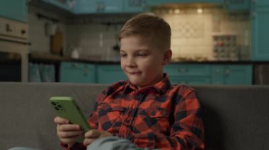 Kid Laughing Using Smartphone. Happy boy holding mobile phone in hands sitting on couch at home. Child watching funny mobile content. 4K