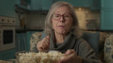 Senior Woman Eating Popcorn at Camera. Mature female looking at camera sitting at home. 4K