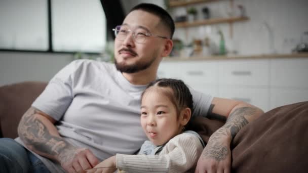 爸爸和女儿看电视吃爆米花 韩国家庭坐在家里的沙发上玩得很开心 可爱的父亲拥抱小女儿 — 图库视频影像