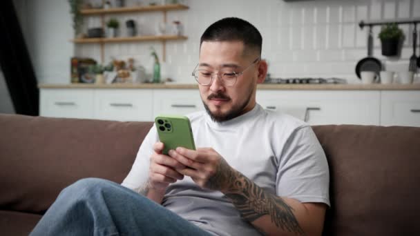 自宅でスマートフォンを使ってテキストを笑顔男 幸せなアジアの男の手のソファの上に座って電話を保持チャット — ストック動画