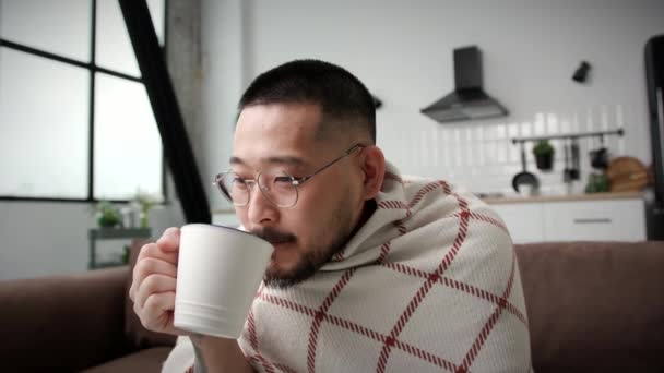 自宅で寒さを感じる30代の男 韓国人男性熱いお茶コーヒーを飲む暖かい取得するために包まれた アパートで暖房がない — ストック動画