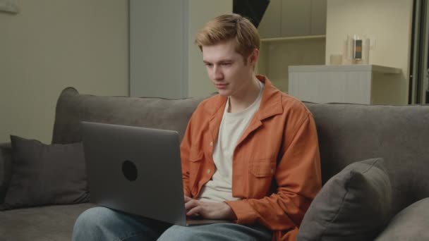 退出的男人在笔记本电脑上阅读好消息 20多岁在网上工作的白人成功地坐在家里的沙发上 — 图库视频影像