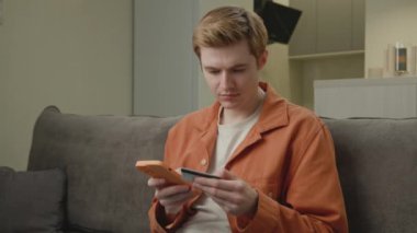 Çevrimiçi Telefon Kullanarak Adam Ödeme. 30 'lu yaşlardaki bir adam banka kredi kartını online alışveriş ve evdeki kanepede oturmak için kullanıyor. 4K