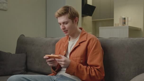 携帯電話を使って20代の男がビデオゲームに勝つ 家庭でスマートフォンをプレイする幸せなゲームの勝者 — ストック動画