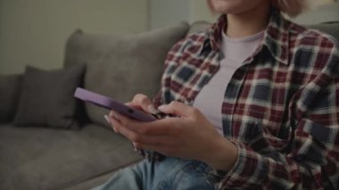 Koltukta oturan cep telefonlu kadın mesajlaşması. Genç yetişkin bir kadın evde akıllı telefonla sohbet ediyor. El kamerasıyla çekilmiş. 4K