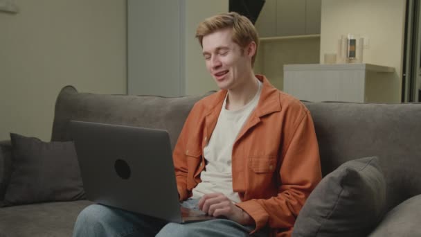 ラップトップを使用したビデオ通話の男 ブロンドの男はソファに座って自宅で働いているコンピュータと話をした — ストック動画