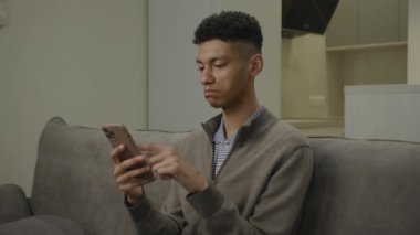 Siyah adam akıllı telefon kullanıyor. Evdeki koltukta oturan Afrikalı Amerikalı adam internette cep telefonu kullanarak sörf yapıyor. 4K