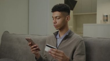 Siyah adam kredi kartı ve cep telefonuyla ödeme yapıyor. Afrikalı Amerikalı erkek online alışveriş yapıyor. Müşteri banka kartıyla ödüyor. 4K