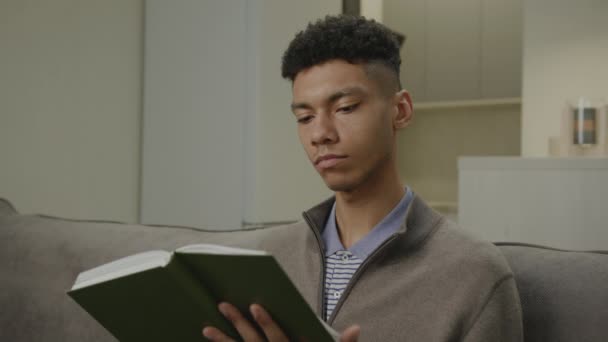 非洲裔美国人在家里读书 千禧年的男性在客厅里看纸质书 对着相机笑 — 图库视频影像