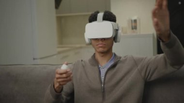 VR 'daki adam elleriyle el hareketi yapıyor. Sanal gerçeklik kulaklığı takan milenyum erkeği evdeki kanepede oturuyor. Sanal gerçeklik yazılıyor. 4K