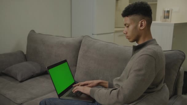 Black Man Πληκτρολογώντας Στο Laptop Πράσινη Οθόνη Κάθεται Στον Καναπέ — Αρχείο Βίντεο