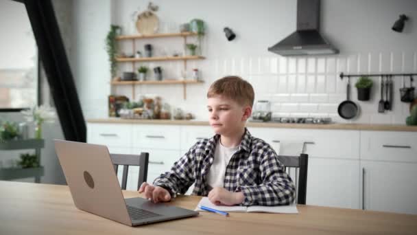学校の少年は自宅でノートパソコンを使ってオンライン学習 ノートに書き オンライン教師に耳を傾ける子供 遠隔教育 — ストック動画