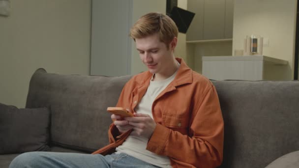 20多岁的男人在沙发上用手机发短信千禧年男性手持智能手机上网浏览 快乐的年轻人在网上冲浪 — 图库视频影像