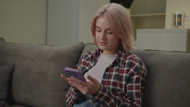 女人在沙发上用电话 20多岁的女士上网浏览和大笑 有趣的内容在线 成年人通过智能手机与朋友聊天 — 图库视频影像