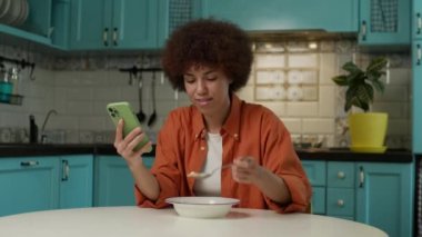 Çevrimiçi Afro-Amerikan Kadın Tarama ve Evde Yeme. Kahvaltıda cep telefonuna bakan bir kadın. 4K