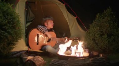 Çocuk Kamp ateşinin yanında Çadırda Oturarak Gitar Çalıyor. Bir çocuk gece ateşin başında şarkı söylüyor. 4K