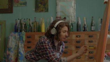 Cheerful Artist Creating Her Masterpiece Listening To Music. Female painter in headphones painting on canvas in workshop. Happy woman working in art space. High quality 4k footage