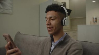 Kulaklıklı Adam Şarkı Söylüyor Cep Telefonu Tutuyor. 20 'li yaşlarda erkek, evdeki koltukta oturan kulaklıkla müzik dinlemekten hoşlanır. 4K
