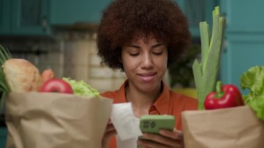 20 'li yaşlardaki kadın market fişini kontrol ediyor. Siyah bir kadın, içi taze yiyecek ve cep telefonuyla dolu market poşetlerine bakıyor. 4K