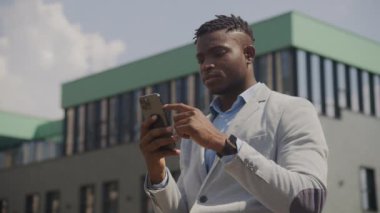 Smartphone Standing Out 'u kullanan siyahi iş adamı. Afro-Amerikan Adam açık havada cep telefonuyla internette geziniyor. 4K