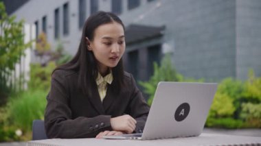 Asyalı İş kadını dışarıdaki bilgisayarı kullanarak görüntülü arama yapıyor. İnternette çalışan 20 'li yaşlarda bir kadın dışarıda oturuyor. 4K