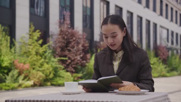 アジアの女性は外で座っている本を読んでいます カフェで本を楽しんでいる若い女性 4Kについて — ストック動画