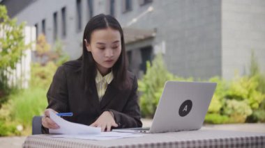 Asyalı iş kadını dizüstü bilgisayar kullanarak belgelerle çalışıyor. Genç bayan girişimci dışarıda kağıtları imzalıyor. 4K