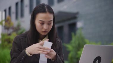 Dışarıdaki finans işleriyle ilgilenen Asyalı iş kadını. Genç yetişkin bir kadın dizüstü bilgisayarla makbuz tutuyor. Kapatın. 4K