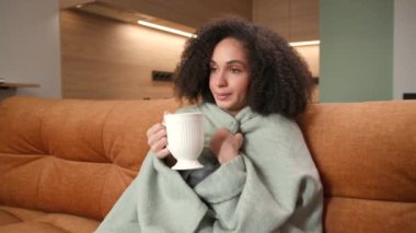 Dairelerde Üşüyen Kadın. Battaniyeye sarılı sıcak çay içen bir kadın kanepede oturuyor. Evde ısıtma eksikliği. 4K
