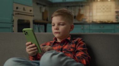 Evde cep telefonu kullanmaktan sıkılmış çocuk. Kanepede tek başına oturan akıllı telefonu kullanarak internette sörf yapan hoşnutsuz çocuk. 4K