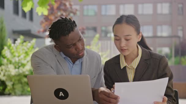 外で働く2人のビジネスマン 黒人男性とアジア人女性はラップトップと文書を使用しています 4Kについて — ストック動画