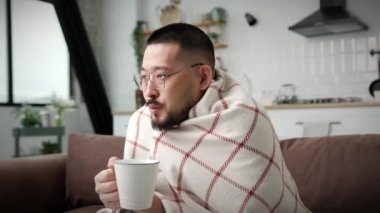 Asyalı adam evde üşüyor. Ekoseye sarılı Koreli adam ısınmak için sıcak çay kahvesi içiyor. Dairelerde ısıtma eksikliği. 4K