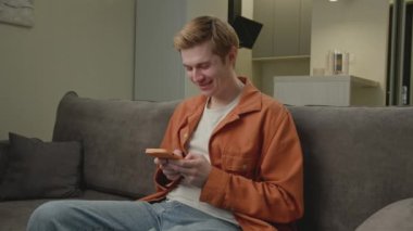 Smartphone 'da gülümseyen adam koltukta oturuyor. Genç yetişkin bir erkek evde akıllı telefondan sohbet ediyor. 4K