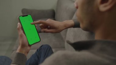 Smartphone Green Screen kullanan adam kanepede dikey oturuyor. Siyah erkek dikey cep telefonu krom anahtarına bakıyor. Omuz manzarasının üstünden. 4K