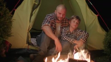 Baba ve Oğul, Kamp Ateşi 'nin yanındaki çadırda oturuyor. Oğlu da babası gibi. Baba ve Kid yumruk tokuşturuyor. 4K