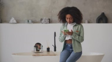 Tuvalette oturan siyah kadın internette cep telefonuyla sörf yapıyor. Afro-Amerikalı kadın akıllı telefondan kayıyor evde dinleniyor. 4K