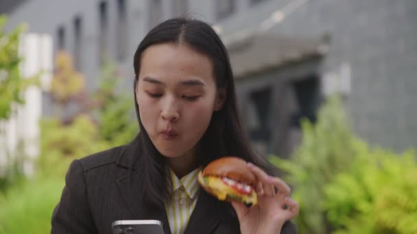 アジアの女性は外でバーガーを食べながら携帯電話をブラウズします カフェでランチを食べ スマートフォンを利用するビジネスマン 4Kについて — ストック動画
