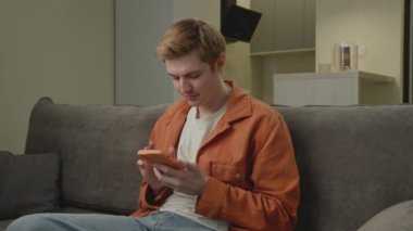 Cep telefonu kullanan adam. 20 'li yaşlarda biri akıllı telefon kullanarak internette sörf yapıyor. Elinde cep telefonu olan genç bir yetişkin kanepeye oturmuş kameraya bakıyor. 4K