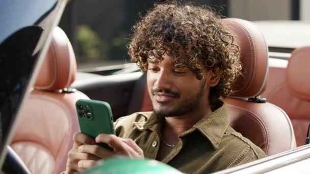 コンバーチブルカーに座っているスマートフォンをスクロールする男 20代の男性が携帯電話でタイピングし カメラを見ながら微笑んでいる 4Kについて — ストック動画