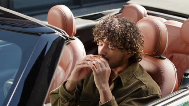 コンバーチブルカーでバーガーを食べる男 車でチーズバーガーを食べているインド人男性の運転手 4Kについて — ストック動画
