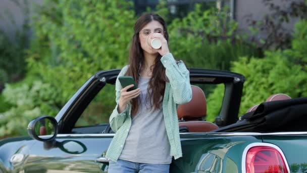 妇女站在汽车附近浏览智能手机喝咖啡 女的在敞篷车附近翻动手机 — 图库视频影像
