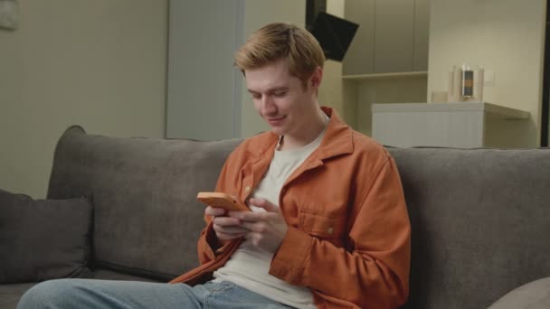 男人用手机 白人用智能手机上网 坐在沙发上 笑着拿着手机的年轻人 — 图库视频影像