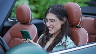 Üstü açık arabada oturan cep telefonundan mesajlaşan kadın sürücü. Arabadaki akıllı telefonlara bakan bir kadın. 4K