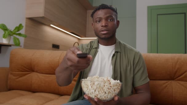 ブラックマンは自宅でカウチに座ってポップコーンを食べます 20代のアフリカ系アメリカ人男性がテレビを見たり カメラを見ているスナックを食べたりします 4Kについて — ストック動画