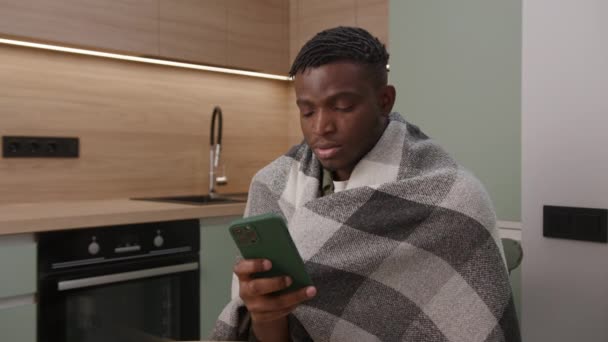 家で寒いと感じている黒人男性 アフリカ系アメリカ人男性が毛布のブラウジング携帯電話で包まれました アパートでの暖房が足りない 4Kについて — ストック動画