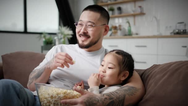 パパと娘はテレビを見てポップコーンを食べる 韓国人の家族は自宅でソファに座って楽しんでいます 愛する父は小さな娘を抱きしめる — ストック動画