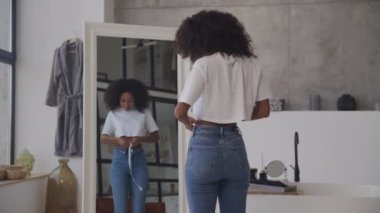 Siyah kadın ölçü bandıyla belini ölçüyor. Aynaya bakarak kilo verdikten sonra Afro-Amerikan kadın. 4K