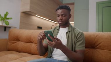 Siyah adam koltukta oturan telefonu kaydırıyor. 20 'li yaşlardaki Afro-Amerikan erkek evde alet bağımlısı. 4K