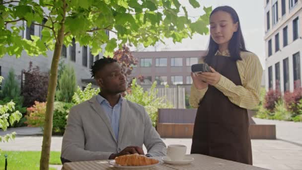 黑人商人与手机和Nfc终端坐在户外咖啡店付款 服务员在餐厅里拿着无接触式付款终端 — 图库视频影像