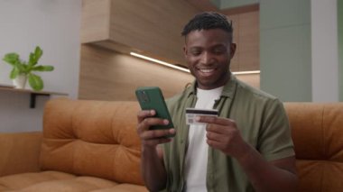 Siyah adam kredi kartıyla ödemeyi telefonla yapıyor. 20 'li yaşlardaki Afro-Amerikan erkek, online alışveriş yapıyor. 4K